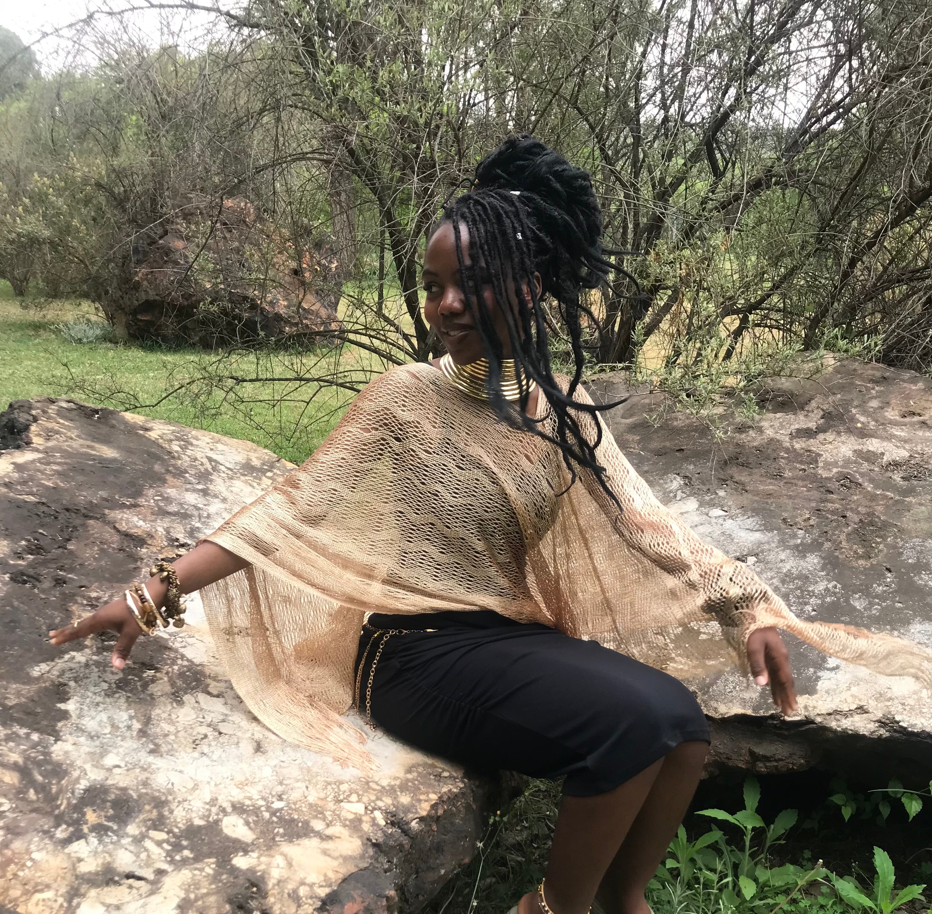 A woman with long black dreadlocks sits on a large rock outdoors, wearing a beige open-knit poncho-style top over a black skirt. Trees and shrubs are visible in the background.